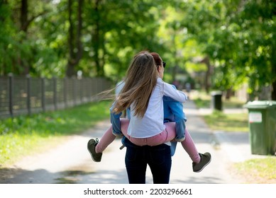 Rear View On Young Daughter On A Piggy Back Ride With Her Mother.Happy Family Having Fun Outdoor