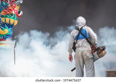 Rear view of outdoor healthcare worker in protective clothing using fogging machine spraying chemical to eliminate mosquitoes and prevent dengue fever at Chinese shrine in Samutsakhon, Thailand