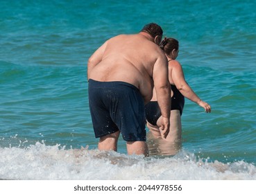 Rear View Of Overweight Couple In The Sea Waves At Shore. Fat Man And Fat Woman On The Beach. Obese Couple Outdoor. Serious Problem With Health