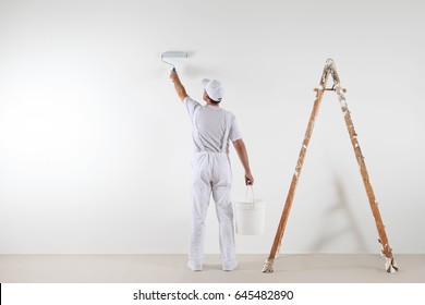 Rear view of painter man painting the wall, with paint roller and bucket, isolated on big empty space with wooden ladder - Shutterstock ID 645482890