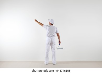 Rear View Of Painter Man Pointing With Finger The Blank Wall, With Paint Roller, Isolated On White Room