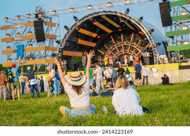 Rear view of people having fun at a summer music festival, enjoying outdoor live performance, with a woman raising her hands. - Powered by Shutterstock