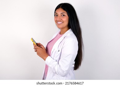 Rear View Photo Portrait Of Young Hispanic Doctor Girl Wearing Coat Over White Background Using Smartphone Smiling