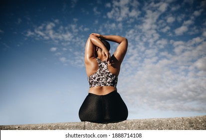 Rear view of a plus size female sitting on wall and stretching outdoors. Female in sportswear relaxing her arm muscles after workout.