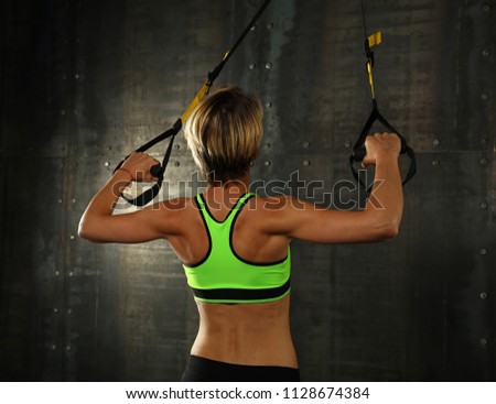 Rear view portrait of one young athletic woman at crossfit training, exercising with trx suspension fitness straps over dark background