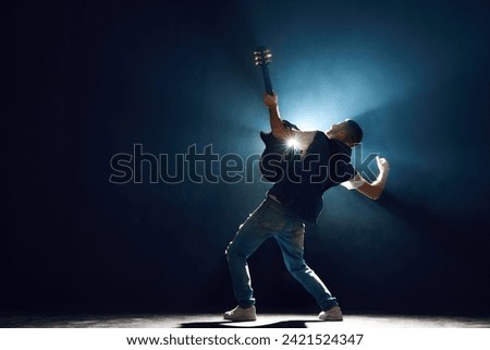 Rear view portrait of street style musician strumming chords on black guitar with dramatic on stage against black background with backlights. Concept of Rock-n-roll, music and dance, culture.