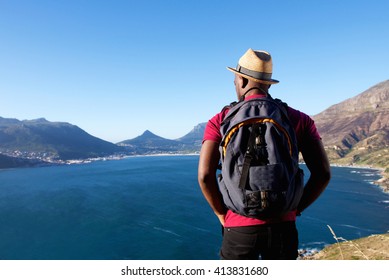 Rear view portrait of young african man on holiday with hat and backpack looking at the sea