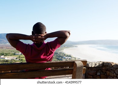 Rear view portrait of young african man sitting relaxed on a bench with his hands behind head, young guy on vacation.