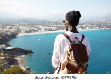 Rear view portrait of young African American backpacker facing sea standing on viewing platform or rock, admiring fascinating views in European resort city, dressed casually. People and travelling