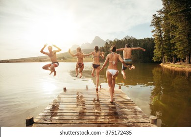 Rear View Portrait Of Young Friends Jumping Into A Lake. Young People Running And Jumping From A Jetty In To A Lake On A Sunny Day.