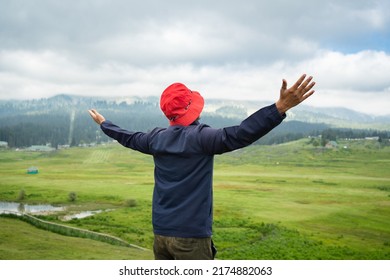 Rear view portrait of young indian guy with red hat and standing outdoors with arms spread open.