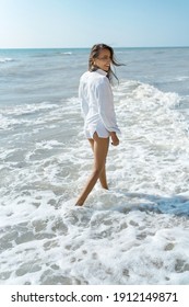 Rear View Positive Latin Woman Walking By Sea Water On Beach At Sunny Day, Wearing Flowy White Shirt. Concept Wellness And Quality Life