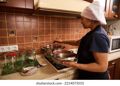 Rear View Of A Pretty Woman, Housewife In Chef's Apron And Black Apron, Standing By A Stove And Cooking Passata From Organic Tomatoes, Making Tomato Sauce For Preserving In Sterilized Cans For Winter