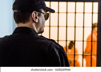 Rear View Of Prison Officer Standing Near Prison Cell With Criminals