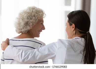 Rear View Profile Smiling Face Of Satisfied Aged Woman Clinic Patient Receiving Support Encouragement From Caring Young Nurse In White Coat, Caregiver Provide Help Hug Old Granny During Visit At Home