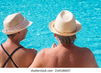 Rear view of relaxed senior couple wearing straw hat in summer time sitting on the edge of the swimming pool. Two happy retirees enjoy their holidays under the sun