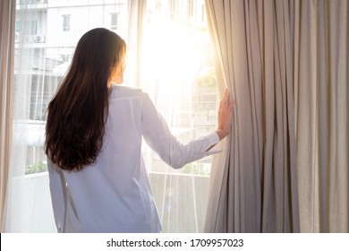 Rear View Of Relaxing Young Beautiful Asian Woman Girl Wake Up In Home Bedroom And Using Her Hand Opening Balcony Window Curtains With Sun Shining Through The Curtain To The Room In Fresh Morning.