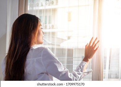 Rear View Of Relaxing Young Beautiful Asian Woman Girl Wake Up In Home Bedroom And Using Her Hand Opening Balcony Window Curtains With Sun Shining Through The Curtain To The Room In Fresh Morning.