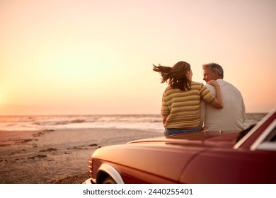 Rear View Of Retired Senior Couple Sitting On Hood Of Classic Sports Car At Beach Watching Sunrise - Powered by Shutterstock