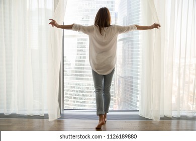 Rear view at rich woman standing looking out of full-length window of luxury modern apartment or hotel room opening curtains in the morning enjoying sunlight and city skyscrapers view feeling happy