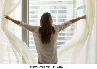 Rear view at rich woman traveler opening window curtains feeling happy free independent, enjoying starting new day, wellbeing, light good pleasant morning of luxury life and big city view concept