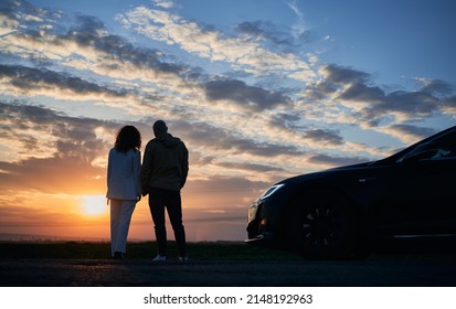 Rear View Of Romantic Couple Who Arrived In Black Car Out Of Town To Admiring Sunset Over Fields At Evening. Pair Of Young People Holding Hand Each Other And Watching Natural Beauty Of Sunset.