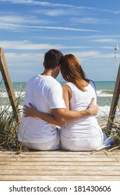 Rear View Of Romantic Man & Woman Couple Sitting On Wooden Steps Overlooking A Beach