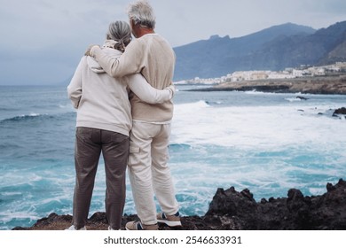 Rear view of a romantic senior couple embracing on a rocky beach on a bad weather day looking waves splashing against the coast enjoying retirement lifestyle or vacation - Powered by Shutterstock