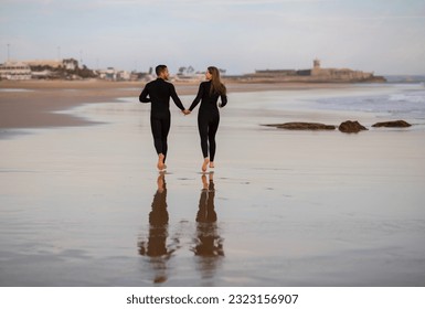 Rear View Of Romantic Young Couple Running On The Beach Together, Happy Loving Millennial Man And Woman Wearing Wetsuits Holding Hands While Walking On Seashore After Surfing, Copy Space - Powered by Shutterstock