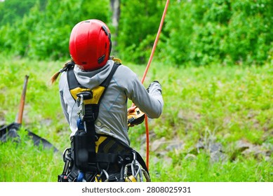 Rear view of a rope access worker in a red helmet descending on an orange rope through a green meadow