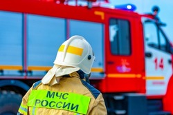 Rear View Of A Russian Firefighter, Rescuer In A Protective Suit And White Helmet Fighting A Fire. Fire Extinguishing Operation. Behind A Sign With The Words "EMERCOM Of Russia"