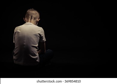 Rear View Of A Sad Silhouette Young Boy Sitting On The Floor Against Black Background With Copy Space.