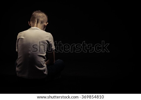 Rear View of a Sad Silhouette Young Boy Sitting on the Floor Against Black Background with Copy Space.