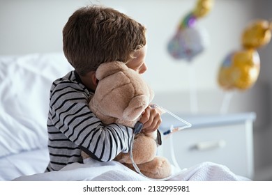 Rear view of scared little boy with intravenous drip in hand hugging teddy bear sitting on hospital bed. Sick lonely child looking through the window in clinic pediatric ward before the surgery.