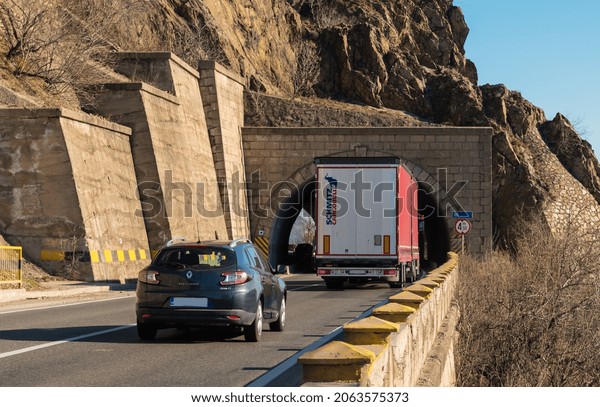 Rear view\
of Schmitz truck with container on the  mountain road, at the\
entrance to the tunnel. Semi-trailer truck carrying goods on\
street. Car in traffic. Italy, October 25,\
2021