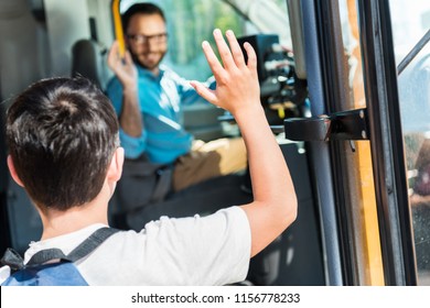 rear view of schoolboy waving to happy bus driver while entering bus