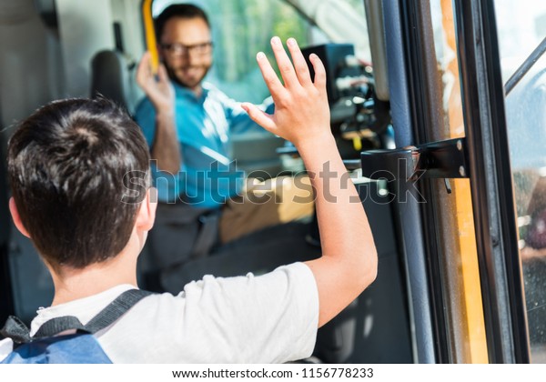 rear view of schoolboy waving to happy bus driver\
while entering bus