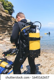 Rear View Of Scuba Diver Man With Oxygen Tank Preparing For Diving . Group Of Divers At The Sea In Background. Summer Extreme Activity .