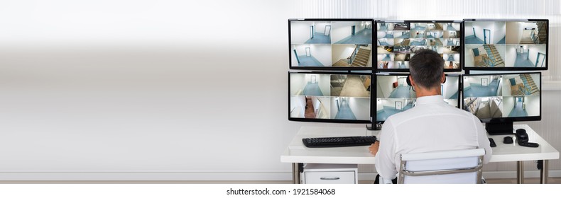 Rear View Of Security System Operator Looking At CCTV Footage At Desk In Office