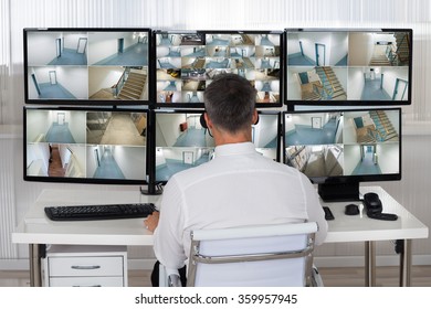 Rear View Of Security System Operator Looking At CCTV Footage At Desk In Office