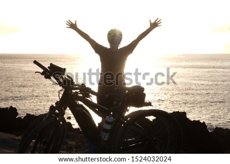 Rear view of senior active woman in front to the ocean at sunset. Black silhouette. Arms raised. Freedon and happiness concept. Yellow helmet.  Electric bici for a healthy lifestyle
