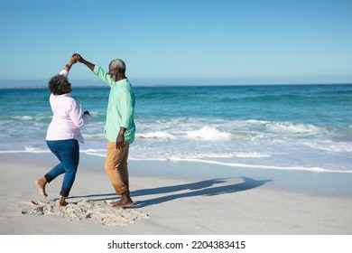 Rear view of a senior African American couple standing on the beach with blue sky and sea in the background, holding hands, dancing and having fun - Powered by Shutterstock