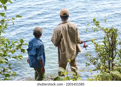 Rear View Of Senior Black Man With Rod And His Grandson Standing In Front Of Lake Or River And Fishing Together While Enjoying Weekend