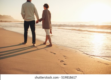 Rear view of a senior couple walking along the sea shore. Mature man and woman together strolling on the sea shore at sunset.