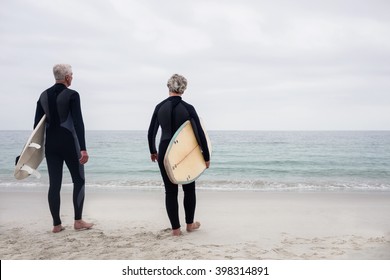 Rear view senior couple in wetsuit holding a surfboard on the beach - Powered by Shutterstock