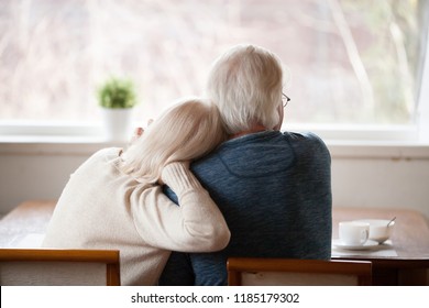 Rear view at senior grey haired loving caring family couple embracing relaxing at home together enjoying peaceful morning breakfast looking at window view thinking of future feeling calm nostalgic