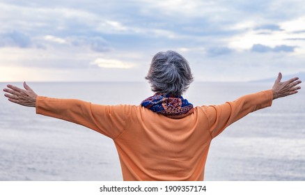 Rear View Of A Senior Grey-haired Woman Enjoying The Freedom And The Horizon Over Water. Standing In Front To The Sea In A Winter Cloudy Day