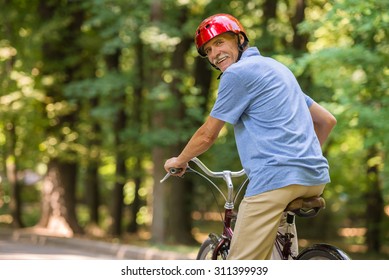 Rear View Of Senior Man In Helmet Is Riding Bicycle In Park And Looking Over Shoulder.