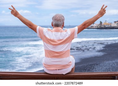 Rear View Of Senior Man Sitting On A Bench Face To The Sea Gesturing Victory With Hands Looking At Horizon Over Water. Active Caucasian Elderly Man  Enjoying Freedom And Vacation