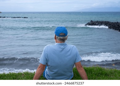 Rear view of senior man sitting on the grass at the beach in a cloudy day enjoying free time vacation or retirement. Relaxed man wearing blue cap looking at the horizon over sea - Powered by Shutterstock